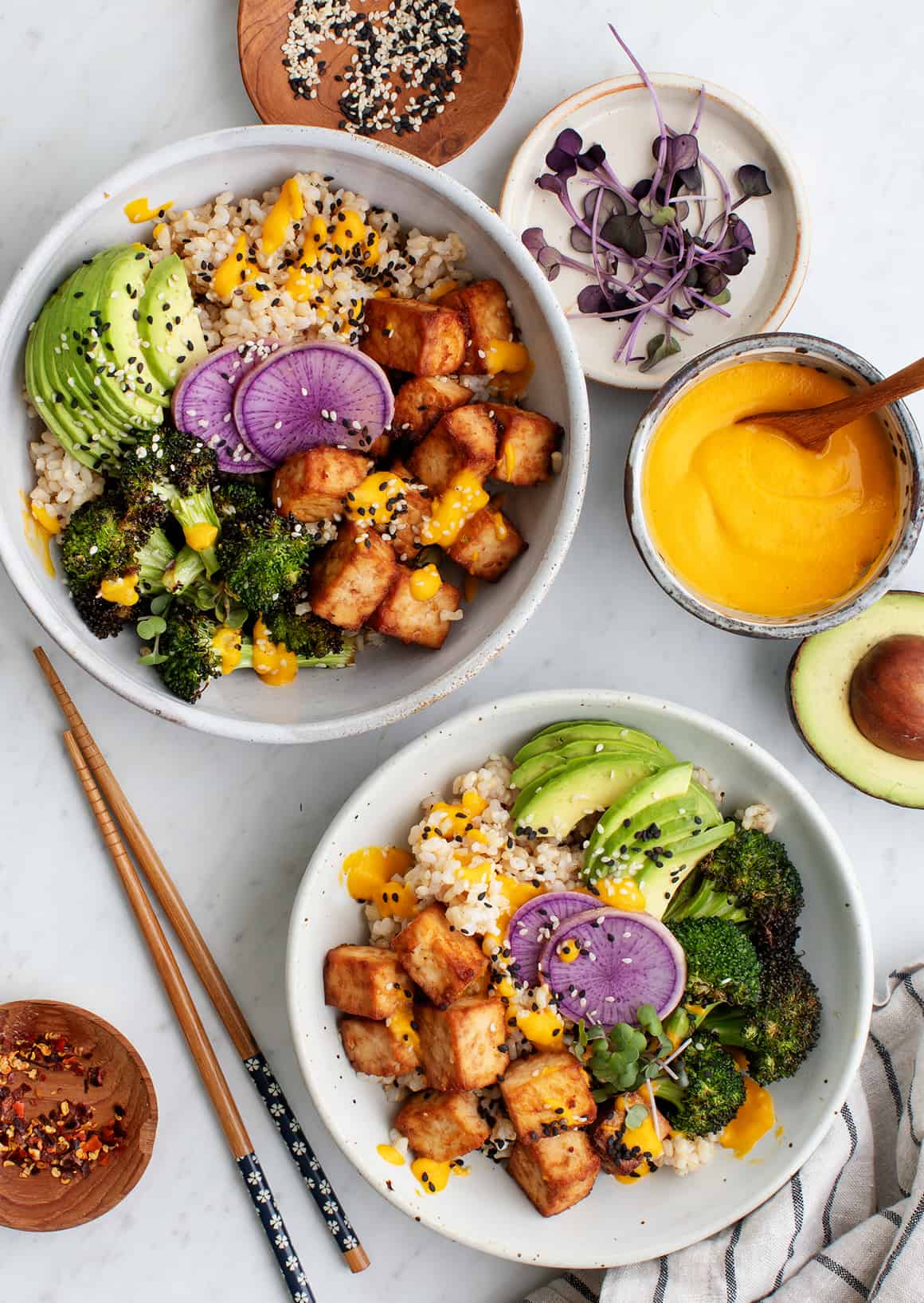 Two bowls filled with brown rice, crispy tofu, broccoli, purple daikon radish slices, avocado, and carrot ginger dressing